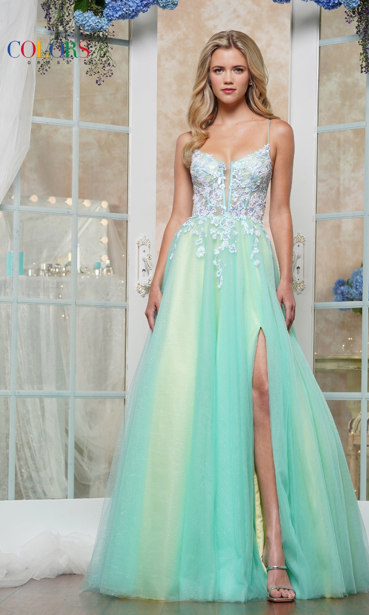 A woman stands before glass doors wearing the Strappy-Back Prom Ball Gown: Colors Dress 3497, a light blue A-line dress with a sheer bodice, floral details, and a high slit.